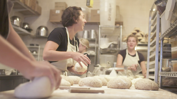 Women stand in a commercial baking kitchen handling loaves of dough