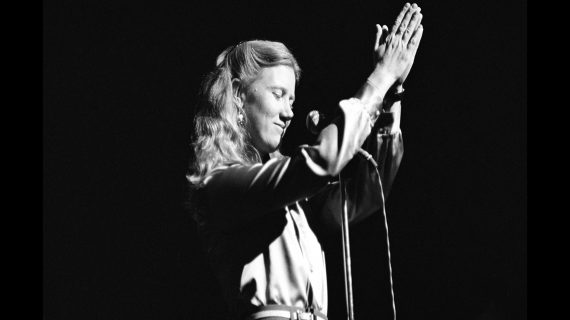 Female singer Holly Near holds her hands in prayer position over her head while standing in front of a microphone.