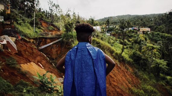 A boy wearing a blue tarp as a cape stands with his back to the camera.