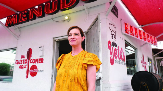 A female chef smiles toward the camera while standing in front of a Mexican food stand.