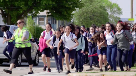 A group of children wearing backpacks run with enthusiasm outside at the close of the school day.