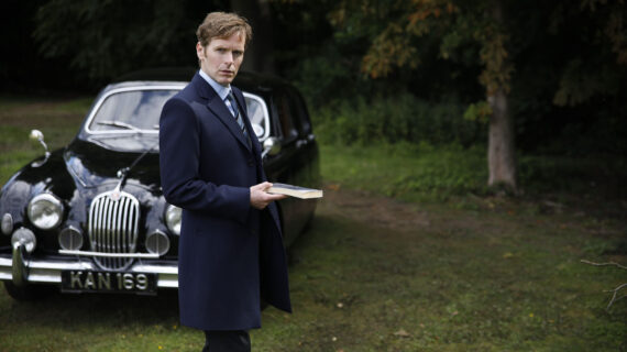 A man in a suit stands in front of an old car.