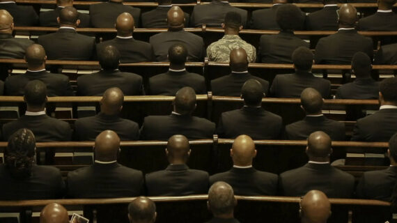 Rows of men in dark suits are seated in church pews, with their backs facing the camera.