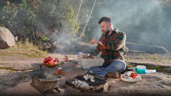 A man sits near an outdoor fire ring cooking fish and apples.