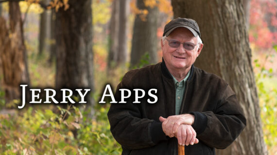 Author Jerry Apps smiling, standing outdoors with autumn trees in the background, leaning on a wooden cane.