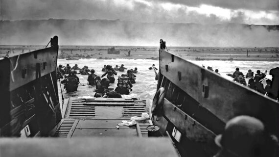 Soldiers wading through the water from a landing craft towards a heavily fortified beach under heavy fire, during a World War II amphibious assault.
