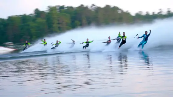 Eight water skiers in a line performing a synchronized routine on a lake, with a forested shoreline in the background.