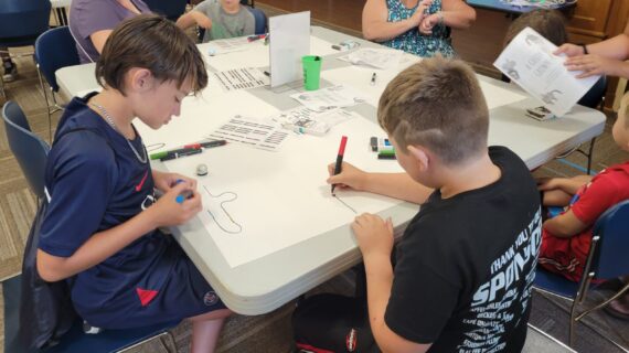 Kids and grown-ups sit at a large table drawing on paper spread across the table.