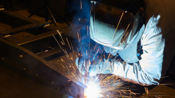 Person wearing a suit and welding mask at work in an industrial setting.