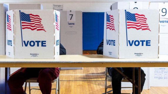 Two voters at a table in a polling place obscured behind cardboard partitions with graphic images of the American flag and the word 