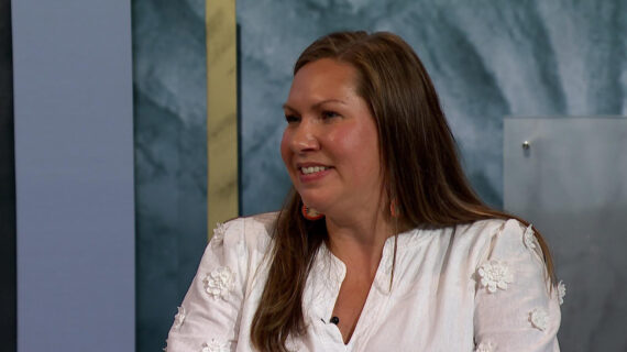 A woman from the shoulders up with long brown hair wears a white blouse and looks to just to her right.