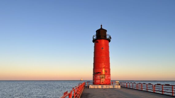 Milwaukee Pierhead Lighthouse at sunset with red fence running along pavement path connecting the lighthouse to the city.