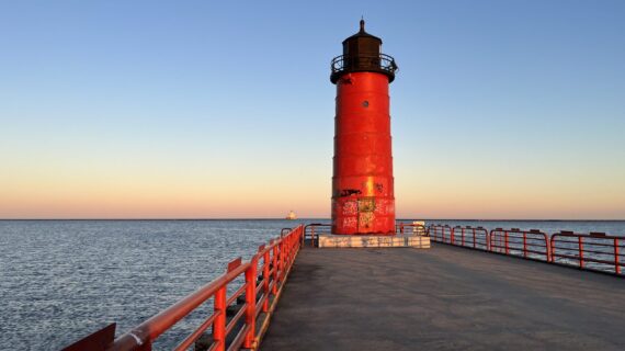 Milwaukee Pierhead Lighthouse at sunset with red fence running along pavement path connecting the lighthouse to the city.