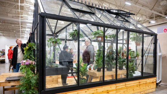 Three people tour a mobile greenhouse display at the 2024 Garden and Landscape Expo with a person inspecting the outside.