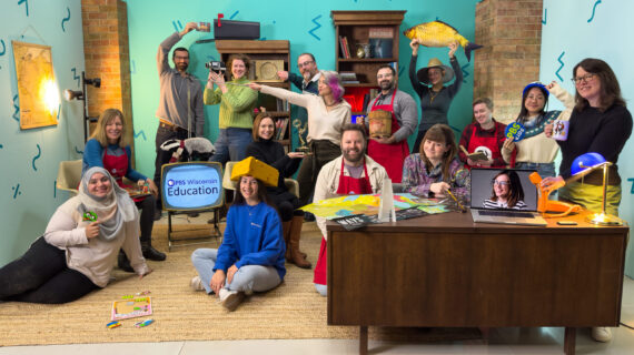 A group of 14 people pose in a playful, colorful office setting surrounded by props, including a cheese hat, toy fish, vintage camera, and PBS Wisconsin Education branding, with one person appearing on a laptop screen.