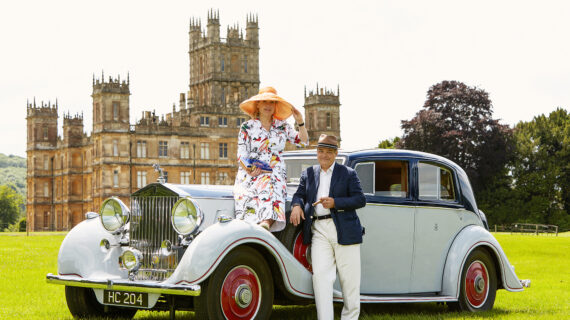 A woman wearing a hat and dress sits on the front of a classic car while her husband, wearing a hat, coat and trousers, stands alongside her holding a cigar. Highclere Castle is in the background.