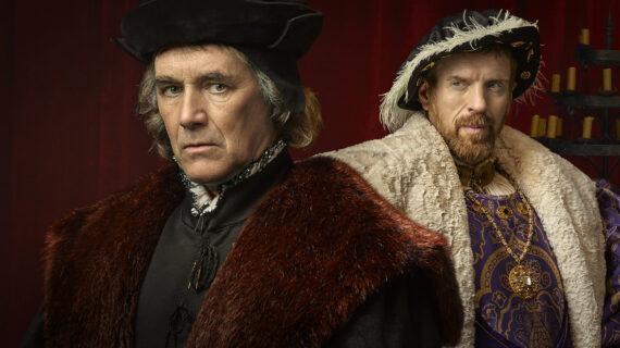 A promotional image from *Wolf Hall: The Mirror and the Light* featuring two men in elaborate Tudor-era clothing. The man in the foreground wears a dark fur-lined coat and a black cap, with a serious expression. The man in the background, dressed in regal purple and gold with a fur cape and feathered hat, looks confidently ahead. The setting is dimly lit with a red curtain and candles in the background, evoking a historical atmosphere.