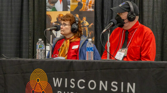 Lisa Johnson (left) and Brian Hudelson (right) sit at a table during WPR's Garden Talk with Larry Meiller live from the 2025 Garden & Green Living Expo with microphones in hand, during an interactive discussion.