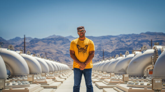 A man wearing a butterfly t-shirt and baseball cap stands in front of a municipal water system with mountains in the background.