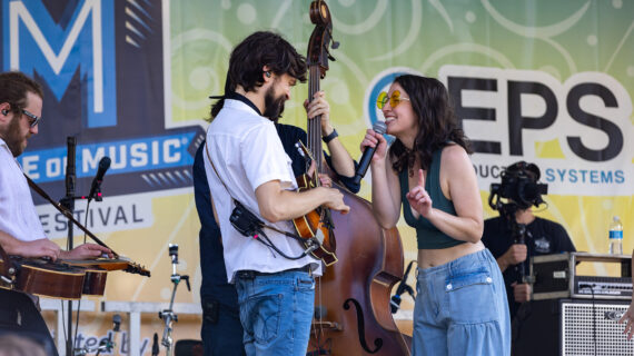 A singer holding a microphone smiles at a guitarist while a cellist plays in the background of an outdoor stage.