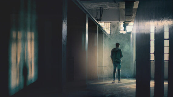 A man dressed in a hooded sweatshirt and jeans stands with his back to the wall in a dimly lit prison enclosure with barred windows.