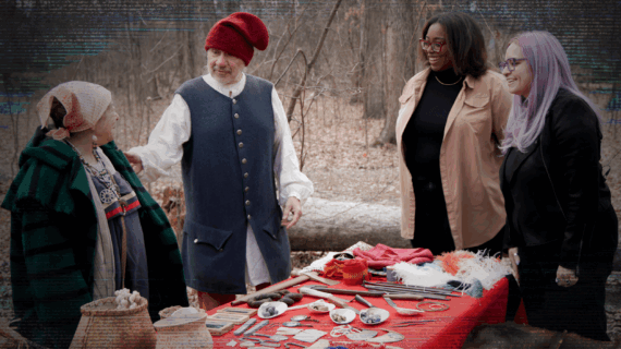 Four people stand around a table outside. The woman and man on the left are dressed in fur trade era clothing. The two women on the right are wearing modern-day clothes.