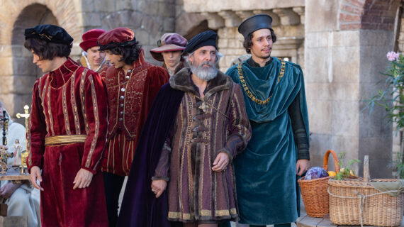 Men in elaborate Renaissance-era costumes stand in a stone courtyard, wearing richly colored velvet garments and hats. One carries a purple cloak and fur-trimmed tunic, while another wears a teal robe with a gold chain. Wicker baskets filled with produce sit on a table nearby.