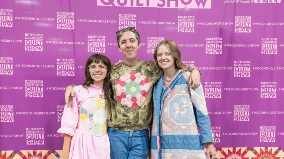 Three young people wearing quilted clothes stand in front of a photo wall with The Great Wisconsin Quilt Show logo on it.