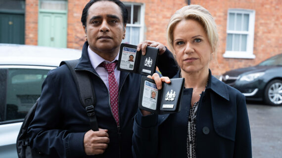 A male and female detective stand side-by-side wearing dark jackets and holding open their identification.