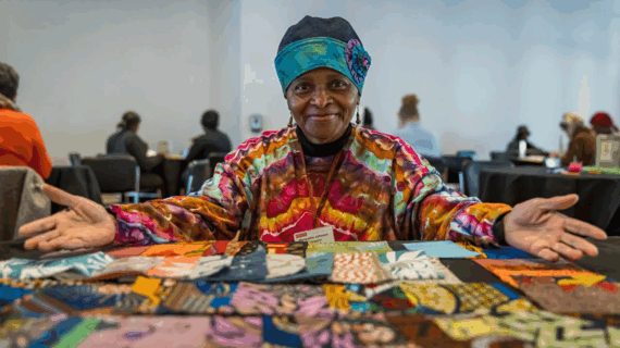 Smiling woman in colorful attire sits at a table displaying vibrant fabric squares, arms open wide, in a room with others working in the background.