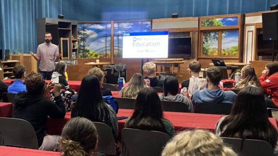Students sit at tables in a media studio, listening to an adult give a presentation.