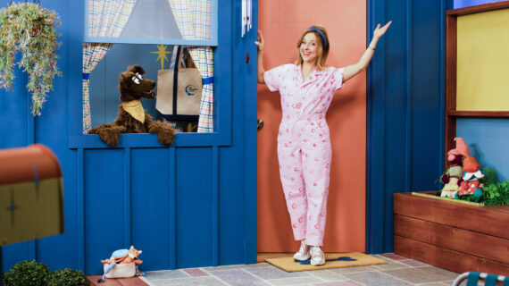 A woman waves at the front door of a blue house. A puppet dog peers out the window at her.