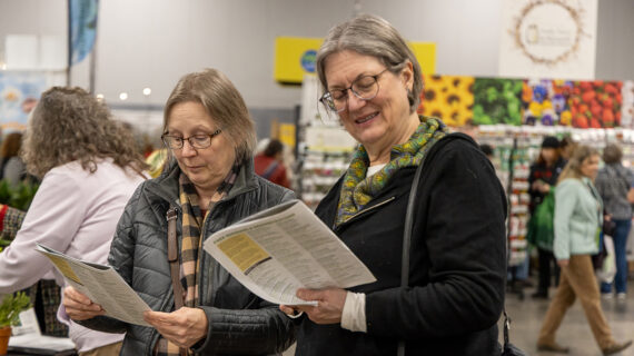 Two people are smiling and reading pamphlets at the 2025 Garden & Green Living Expo at the Alliant Energy Coliseum in Madison, Wisconsin. They are dressed warmly, surrounded by people and colorful displays.