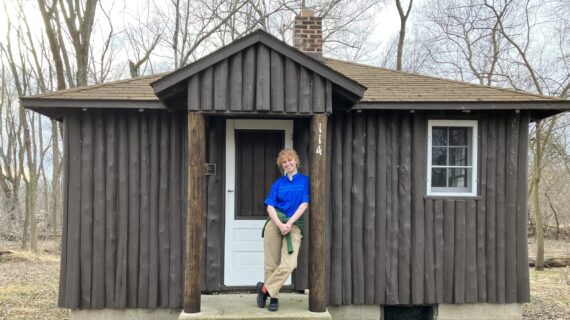 PBS Wisconsin archivist Clara Wolfe poses in front of a quaint cabin, with trees and nature in the background.