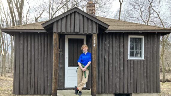 PBS Wisconsin archivist Clara Wolfe poses in front of a quaint cabin, with trees and nature in the background.