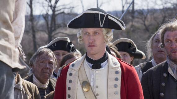 A stern man in an 18th-century British military uniform with a tricorne hat and white wig stands in front of a group of people, some in colonial attire, outdoors on a cloudy day.
