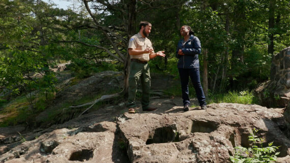 Two people stand on large rocks surrounded by lush greenery and trees. One appears to be a park ranger. The scene conveys an educational tone.