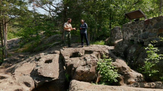Two people stand on large rocks surrounded by lush greenery and trees. One appears to be a park ranger. The scene conveys an educational tone.