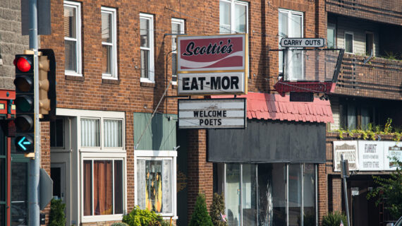 A brick downtown building with a restaurant sign reading “Scottie’s Eat-Mor.” Below it, a marquee says “WELCOME POETS.” An old-fashioned arrow sign above points to “CARRY OUTS.” A traffic light with a green left arrow is visible in the foreground.
