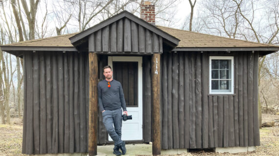 A man stands leaning against the doorway of a small dark brown log cabin with a white door and window, holding a camera in one hand. Bare trees surround the cabin on an overcast day.