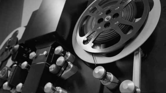 Close-up view of a 16mm film scanner with reels of film threaded through rollers and spools, shown in black and white.