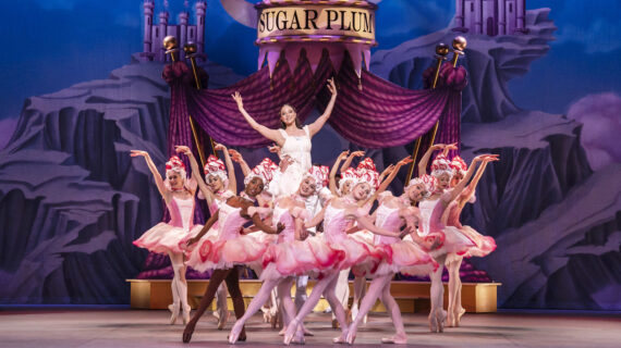 A large group of ballerinas dressed in pink perform on stage in front of a backdrop that includes a curtain and Sugar Plum sign.