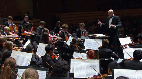 A man conducts a high school orchestra in formal attire at a concert setting.
