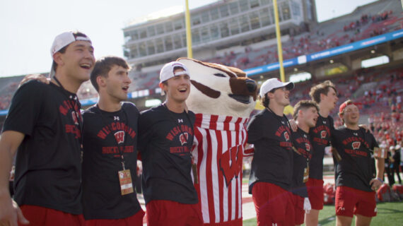 Seven people in matching Wisconsin Badgers gear cheerfully pose on the football field at Camp Randall Stadium in Madison, Wisconsin with the Bucky Badger mascot in red and white stripes, conveying joy and team spirit. Cecil Powless is immediately to the left of the Bucky Badger.