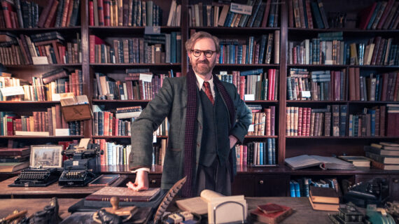 A man in a gray three-piece suit, glasses and scarf stares at the camera while standing at a desk and against a backdrop of bookshelves overflowing with books.