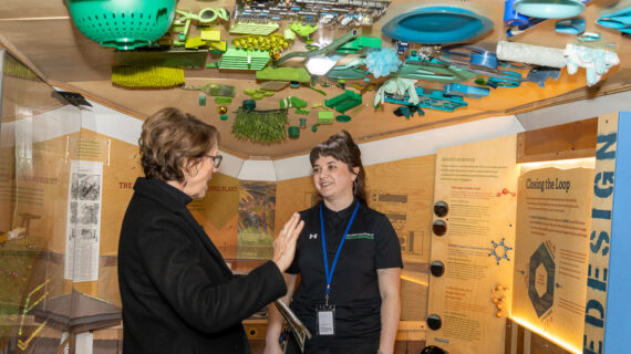 Dane County Department of Waste & Renewables Hanna Kohn (right) in conversation with a visitor inside the Dane County Trash Lab in an exhibit featuring eco-friendly designs at the 2025 Garden & Green Living Expo. The ceiling is adorned with green and blue recycled items.