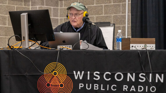 Garden Talk show host Larry Meiller wearing headphones sits at a desk with recording equipment and a computer, behind a table with a Wisconsin Public Radio banner.