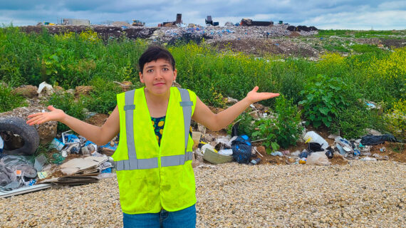 A woman wearing a hi-vis vest standing in front of a landfill with her arms outstretched and shoulders shrugged.