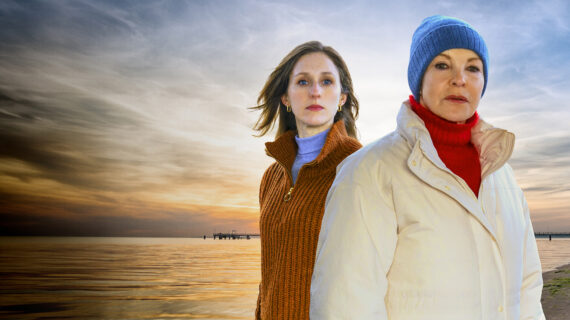 Two women dressed in winter sweaters stand before a sunset over water.