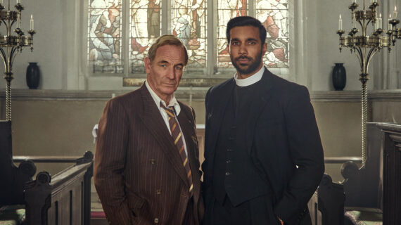 A man wearing a brown suit and tie stands alongside a vicar inside a church. In the background, sunlight shines through a stained glass window.